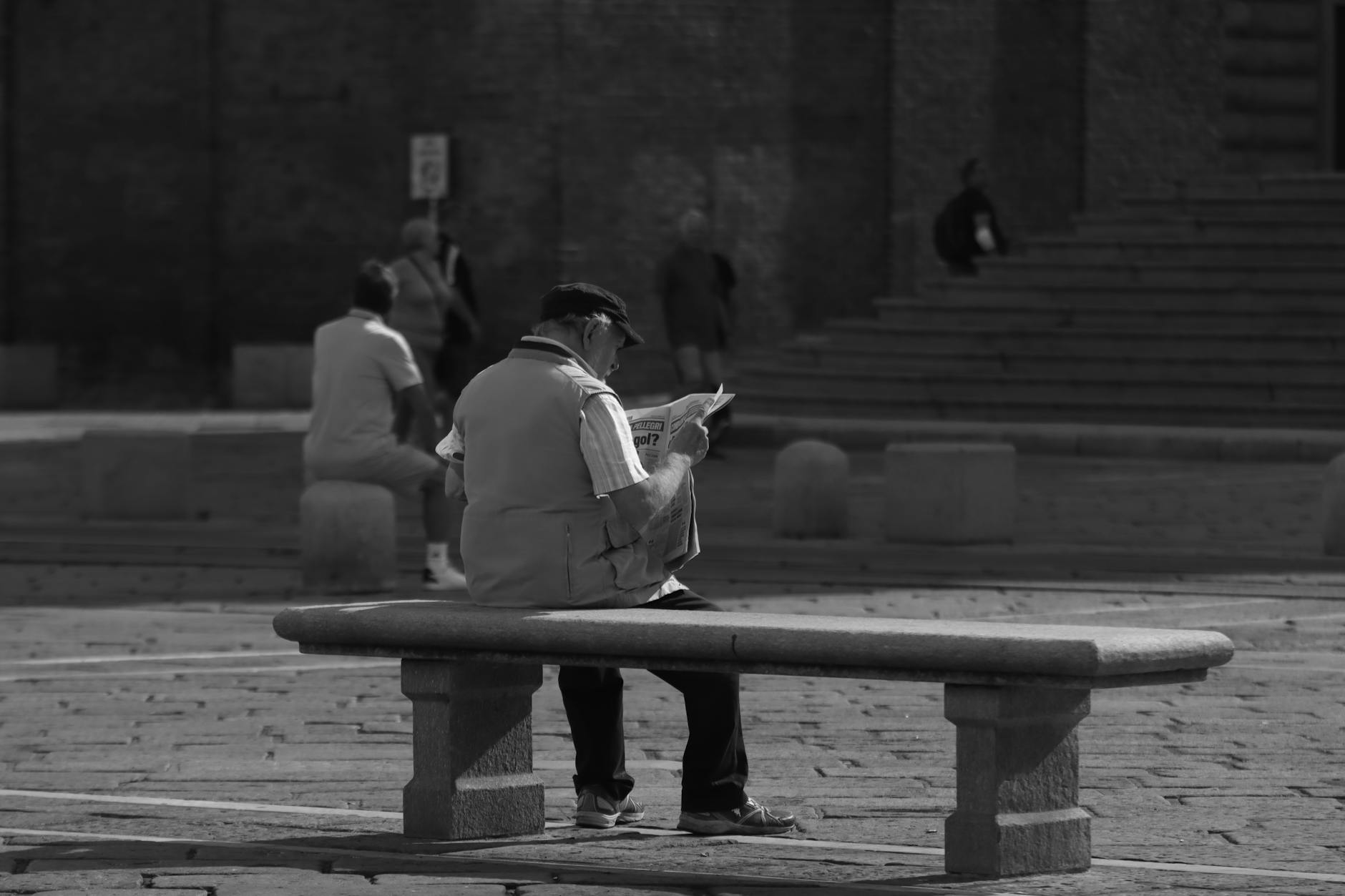 Black and white photo of a senior man reading a newspaper on a stone bench in a plaza.