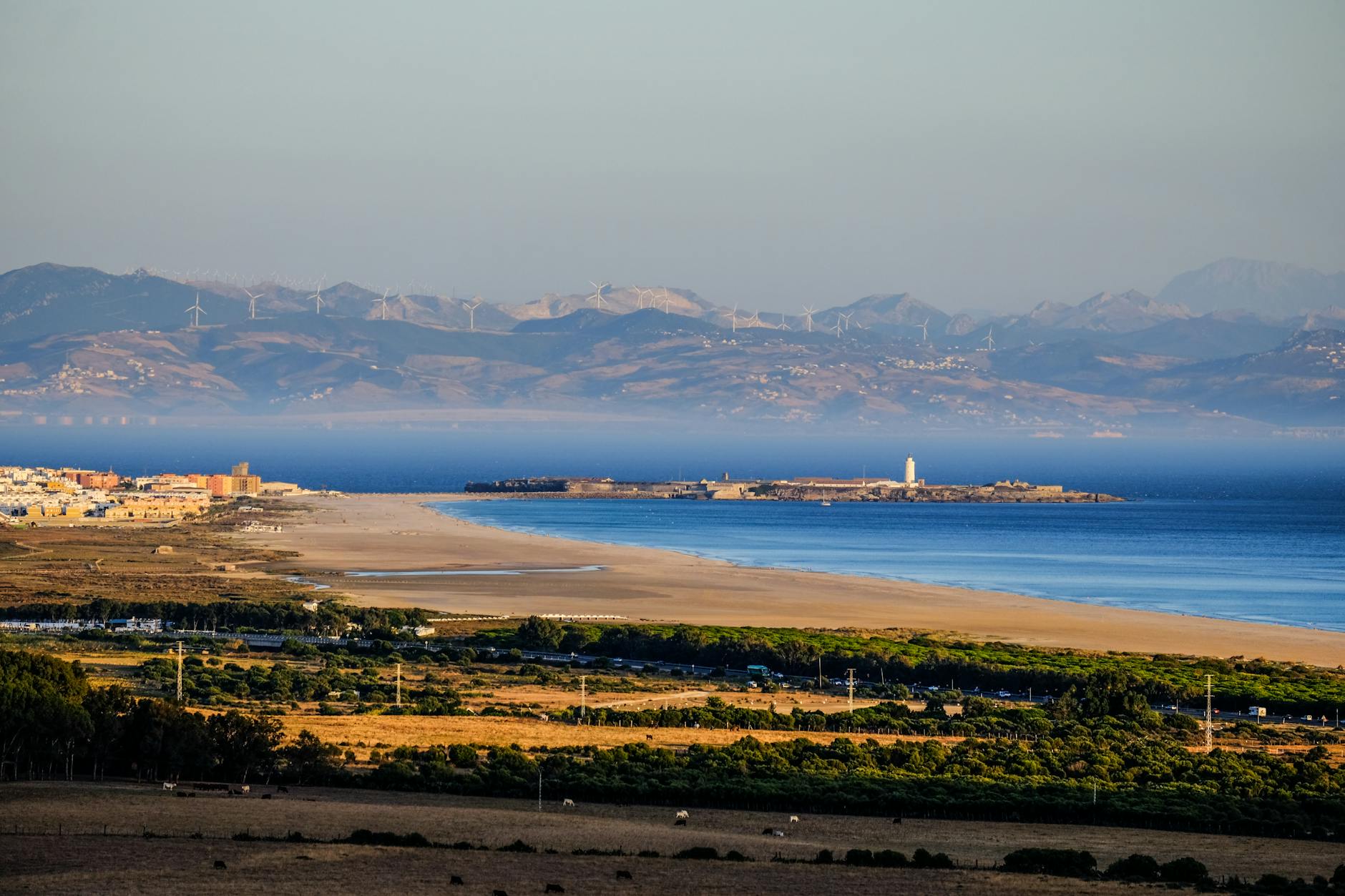 Breathtaking coastal landscape featuring a lighthouse, windmills, and distant mountains under a clear sky.