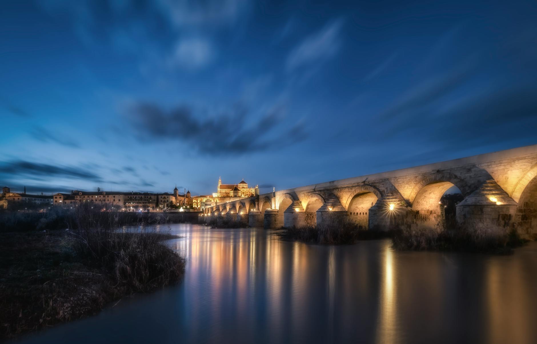 Captured at night, the illuminated Roman bridge in Córdoba spans the tranquil river, reflecting historic architecture.
