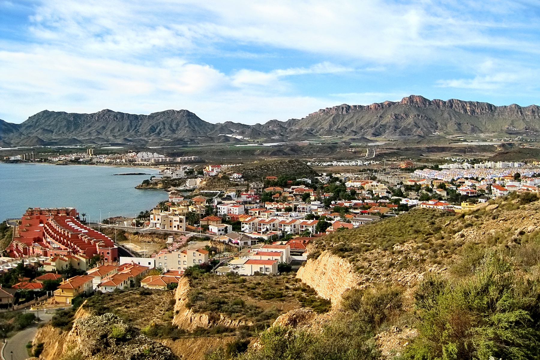 Panorámica diurna que combine la costa del levante almeriense y el entorno de Pulpí: primer plano de una playa del Levante (olas suaves), al fondo el perfil de San Juan de los Terreros y, en una in…
